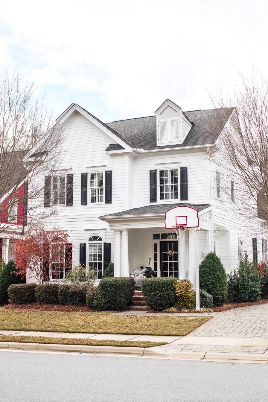 Traditional two-story white house exterior with black shutters, porch columns, a dormer window, and landscaping, in a suburban setting.